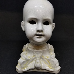 Puede incluir: Cabeza de mu&ntilde;eca de cer&aacute;mica blanca con efecto craquelado. La cabeza tiene cuencas de ojos vac&iacute;as y la boca ligeramente abierta. Est&aacute; montada sobre una base decorativa con motivos florales y de hojas, y detalles amarillos.
