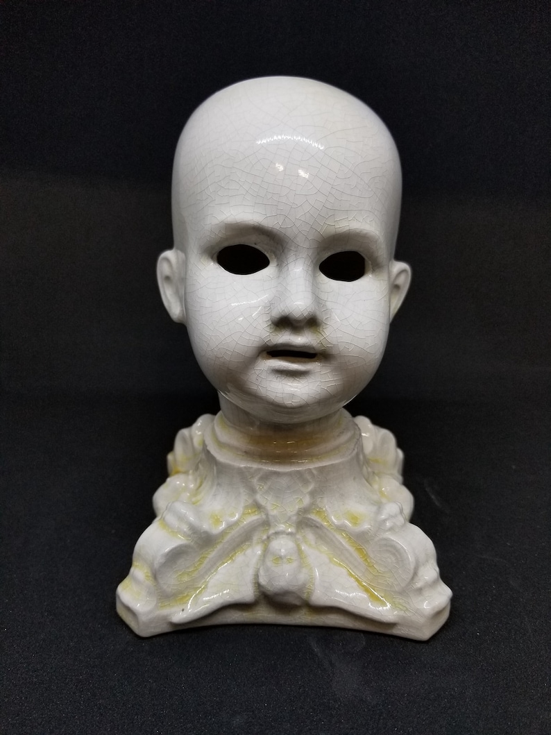Puede incluir: Cabeza de mu&ntilde;eca de cer&aacute;mica blanca con efecto craquelado. La cabeza tiene cuencas de ojos vac&iacute;as y la boca ligeramente abierta. Est&aacute; montada sobre una base decorativa con motivos florales y de hojas, y detalles amarillos.