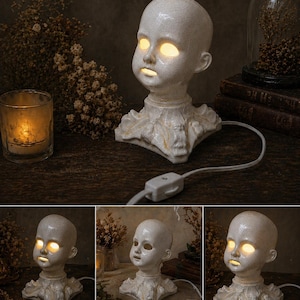 Puede incluir: L&aacute;mpara de cabeza de mu&ntilde;eca de cer&aacute;mica blanca con ojos y boca amarillos brillantes. La l&aacute;mpara tiene un acabado craquelado y se asienta sobre una base decorativa. Un cable blanco con un interruptor de encendido/apagado se extiende desde la base.