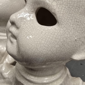 Puede incluir: Primer plano de una cabeza de mu&ntilde;eca de cer&aacute;mica blanca con un acabado agrietado. La cabeza de la mu&ntilde;eca tiene grandes cuencas de los ojos y una cara detallada. La cer&aacute;mica tiene un acabado brillante. La cabeza de la mu&ntilde;eca es un objeto decorativo.