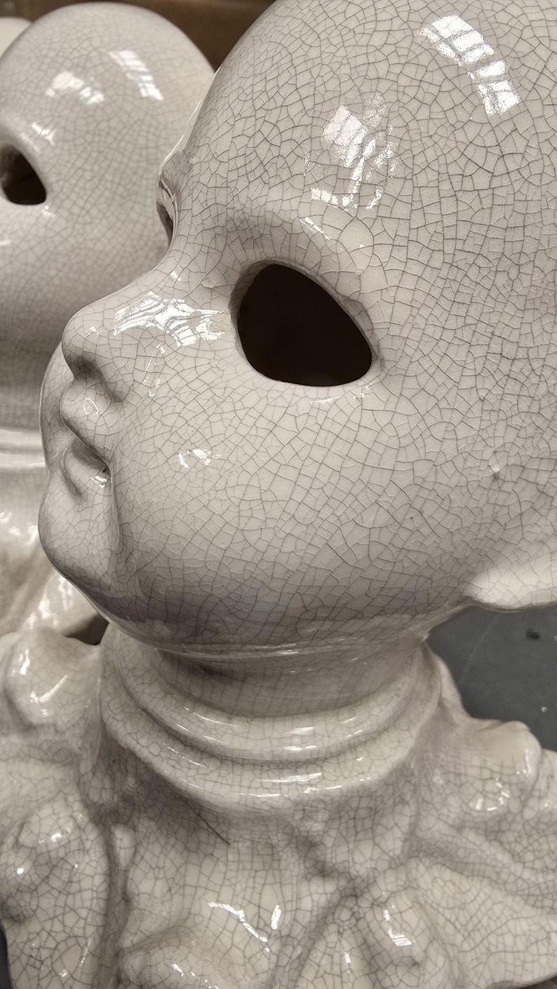 Puede incluir: Primer plano de una cabeza de mu&ntilde;eca de cer&aacute;mica blanca con un acabado agrietado. La cabeza de la mu&ntilde;eca tiene grandes cuencas de los ojos y una cara detallada. La cer&aacute;mica tiene un acabado brillante. La cabeza de la mu&ntilde;eca es un objeto decorativo.