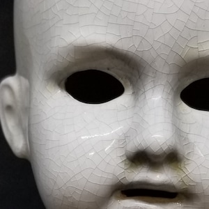 Puede incluir: Primer plano de una cabeza de mu&ntilde;eca de cer&aacute;mica blanca con un aspecto agrietado y envejecido. La cara de la mu&ntilde;eca presenta cuencas de ojos vac&iacute;as, una nariz peque&ntilde;a y una boca ligeramente abierta. La superficie tiene una red de finas grietas.