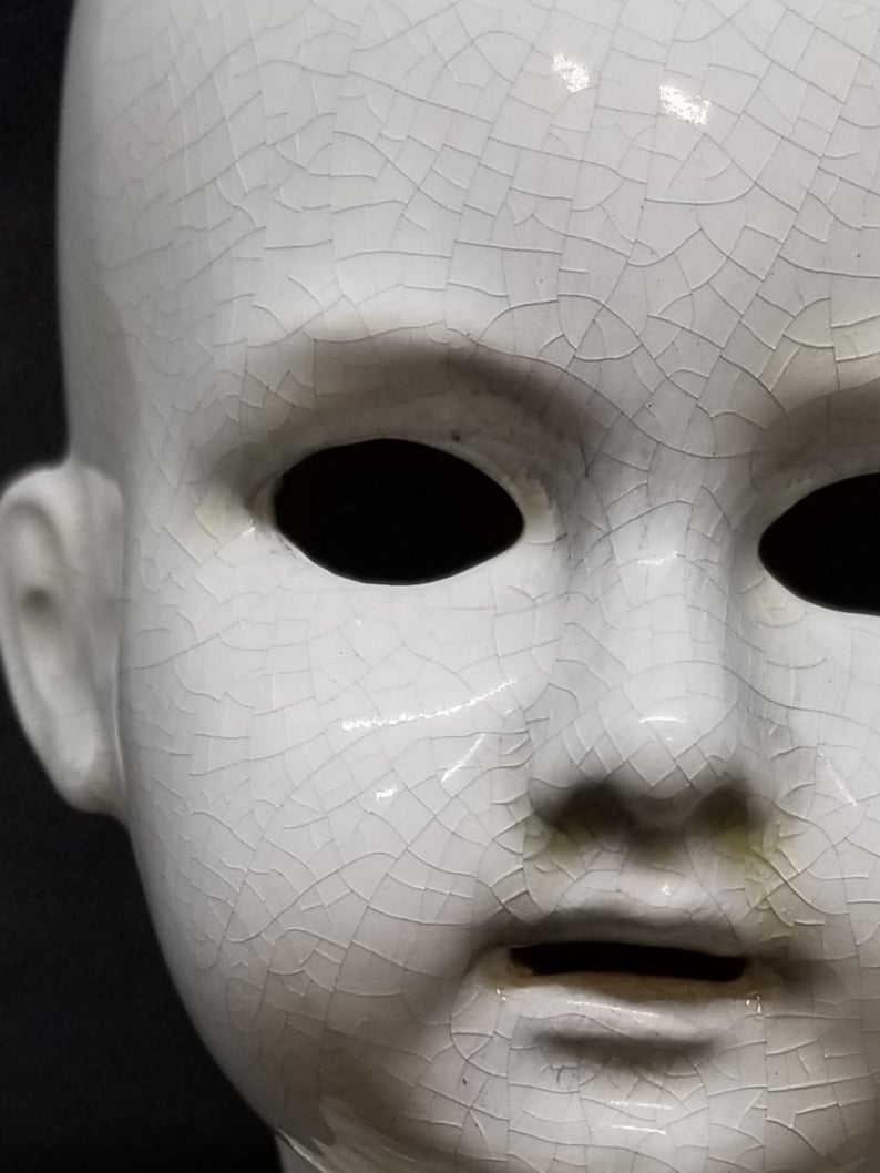 Puede incluir: Primer plano de una cabeza de mu&ntilde;eca de cer&aacute;mica blanca con un aspecto agrietado y envejecido. La cara de la mu&ntilde;eca presenta cuencas de ojos vac&iacute;as, una nariz peque&ntilde;a y una boca ligeramente abierta. La superficie tiene una red de finas grietas.