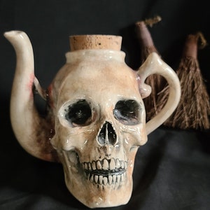 May include: A ceramic teapot shaped like a skull with a cork stopper. The teapot is painted in shades of white, brown, and black, with details of the skull's features.