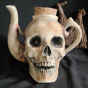 May include: A ceramic teapot shaped like a human skull, with a cork stopper. The skull-shaped teapot is off-white with brown and red accents, and a cracked glaze finish. The handle and spout are curved. The background is black.