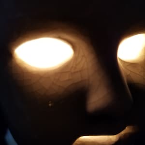 Puede incluir: Una m&aacute;scara de cer&aacute;mica con ojos y boca blancos brillantes, sobre un fondo oscuro. La m&aacute;scara tiene una superficie agrietada y texturizada, y la luz crea un efecto dram&aacute;tico y misterioso. El tema general es misterioso y art&iacute;stico.