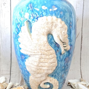 May include: A tall, ceramic vase with a blue and turquoise glaze. A raised, off-white seahorse design adorns the front. The vase is surrounded by seashells and sand, suggesting a coastal theme. The vase is approximately 12 inches tall.