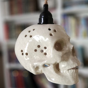 May include: A ceramic skull pendant light fixture with a black fixture and a woven brown cord. The skull is off-white with small holes and a cracked glaze finish. The light fixture is a unique home decor item.
