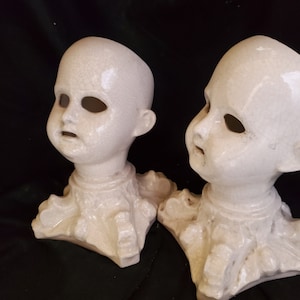 Puede incluir: Dos cabezas de mu&ntilde;eca antiguas de cer&aacute;mica blanca con cuencas de ojos y bocas abiertas. Las cabezas est&aacute;n unidas a una base decorativa. La cer&aacute;mica tiene un aspecto agrietado y envejecido. El fondo es oscuro.
