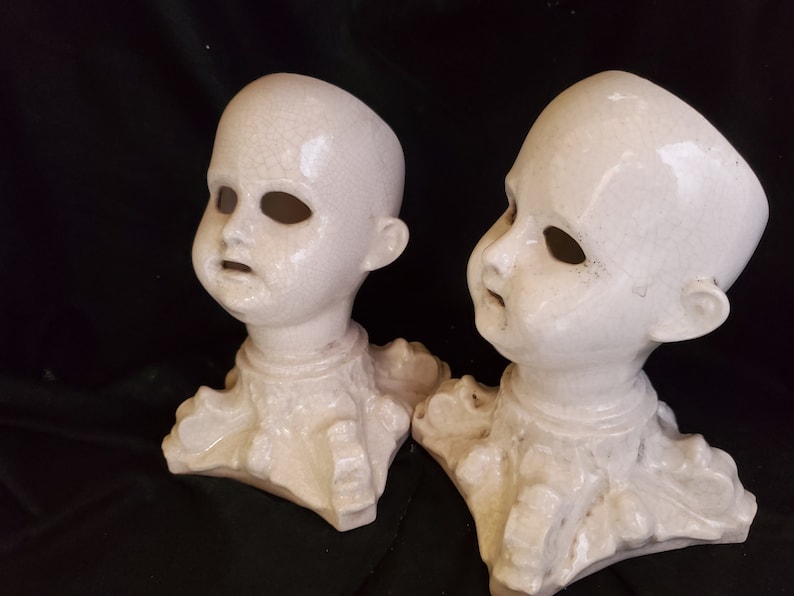 Puede incluir: Dos cabezas de mu&ntilde;eca antiguas de cer&aacute;mica blanca con cuencas de ojos y bocas abiertas. Las cabezas est&aacute;n unidas a una base decorativa. La cer&aacute;mica tiene un aspecto agrietado y envejecido. El fondo es oscuro.