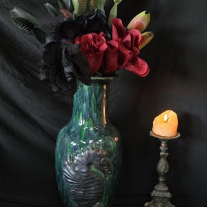May include: A tall, green and blue glazed vase with a seahorse design, filled with black and red artificial flowers and Venus flytraps. A lit candle sits in a decorative metal candlestick. The background is a dark fabric.