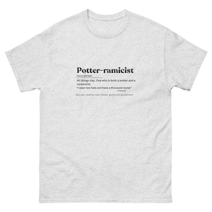May include: A light gray t-shirt with the word "Potter-ramicist" printed in black, along with a definition. The definition describes a person who is both a potter and a ceramicist. The shirt also includes a quote and related terms.