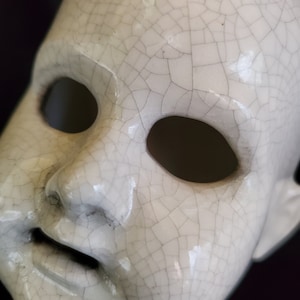 Puede incluir: Primer plano de una cara de mu&ntilde;eca de cer&aacute;mica blanca con grandes cuencas de ojos vac&iacute;as y una boca ligeramente abierta. La superficie est&aacute; cubierta por una red de finas grietas, lo que le da una apariencia envejecida. Los rasgos de la mu&ntilde;eca est&aacute;n sutilmente definidos.