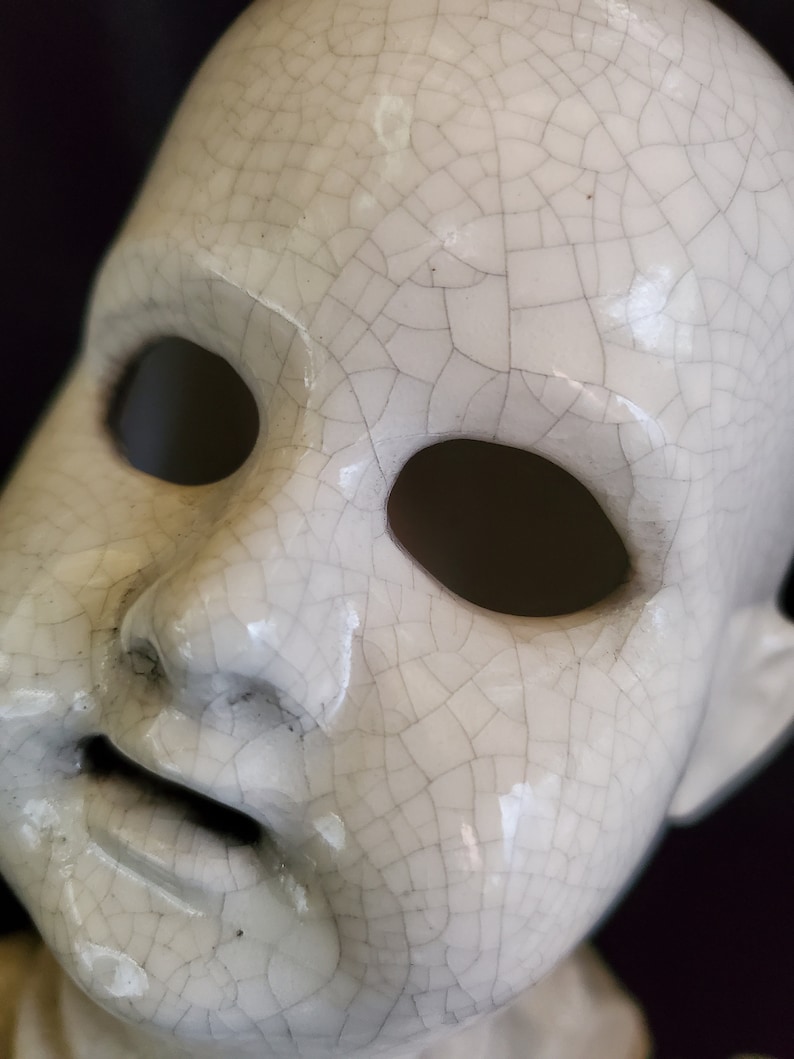 Puede incluir: Primer plano de una cara de mu&ntilde;eca de cer&aacute;mica blanca con grandes cuencas de ojos vac&iacute;as y una boca ligeramente abierta. La superficie est&aacute; cubierta por una red de finas grietas, lo que le da una apariencia envejecida. Los rasgos de la mu&ntilde;eca est&aacute;n sutilmente definidos.