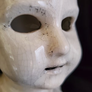 Puede incluir: Primer plano de una cabeza de mu&ntilde;eca de cer&aacute;mica blanca con un acabado de esmalte agrietado. La mu&ntilde;eca tiene cuencas de ojos vac&iacute;as y una boca peque&ntilde;a y abierta. Las motas negras est&aacute;n dispersas por la superficie, lo que se suma a la apariencia envejecida. El fondo es oscuro.