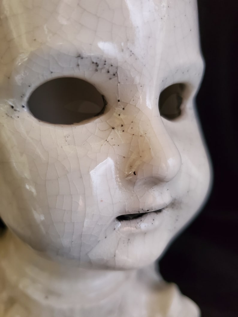 Puede incluir: Primer plano de una cabeza de mu&ntilde;eca de cer&aacute;mica blanca con un acabado de esmalte agrietado. La mu&ntilde;eca tiene cuencas de ojos vac&iacute;as y una boca peque&ntilde;a y abierta. Las motas negras est&aacute;n dispersas por la superficie, lo que se suma a la apariencia envejecida. El fondo es oscuro.