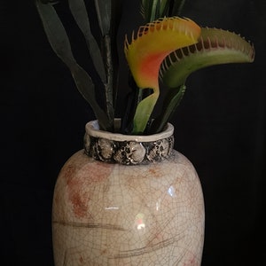 May include: A decorative vase with a crackle glaze finish, holding artificial Venus flytraps. The vase is off-white with red accents and a skull-decorated rim. The flytraps have green and orange-red leaves.