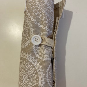 May include: A beige fabric pencil roll with a white button closure and a white mandala pattern. The roll is rolled up and tied with a beige ribbon.