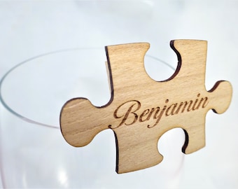 May include: A wooden puzzle piece with the name "Benjamin" engraved in a cursive font. The light-colored wood piece is positioned over a clear glass, suggesting it's a place card or decorative item for an event.