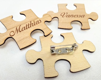 May include: Wooden puzzle piece name tags with the names "Matthias" and "Vanessa" engraved on them. The name tags are light brown and have a pin on the back. The puzzle pieces are scattered on a white surface.