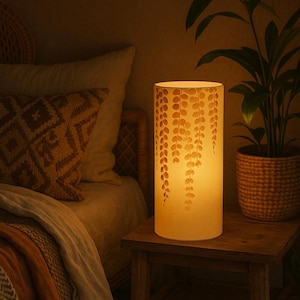 Puede incluir: Lámpara de mesa cilíndrica con luz cálida. La pantalla crema presenta un diseño de hojas recortadas. Se encuentra sobre una pequeña mesita de noche de madera, junto a una cama con cojines decorativos y una planta en una cesta tejida. La lámpara mide unos 30 cm de alto.