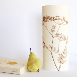 K&ouml;nnte beinhalten: Ein wei&szlig;er zylindrischer Lampenschirm mit braunem Blumendesign. Der Lampenschirm steht auf einer wei&szlig;en Oberfl&auml;che neben einem Buch und einer gr&uuml;nen Birne.