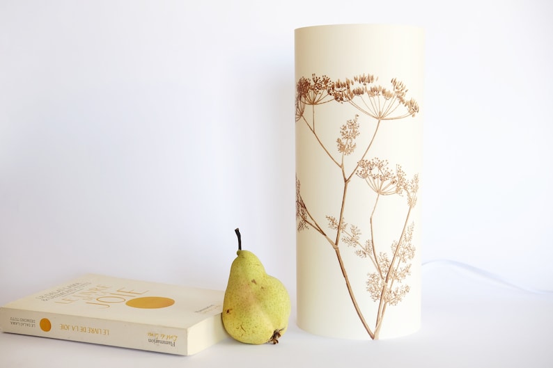 K&ouml;nnte beinhalten: Ein wei&szlig;er zylindrischer Lampenschirm mit braunem Blumendesign. Der Lampenschirm steht auf einer wei&szlig;en Oberfl&auml;che neben einem Buch und einer gr&uuml;nen Birne.