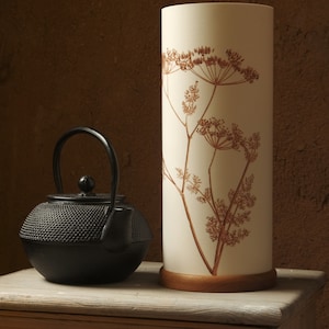 K&ouml;nnte beinhalten: Eine schwarze gusseiserne Teekanne mit Henkel steht auf einem Holztisch neben einer wei&szlig;en Lampe mit braunem Blumendesign.