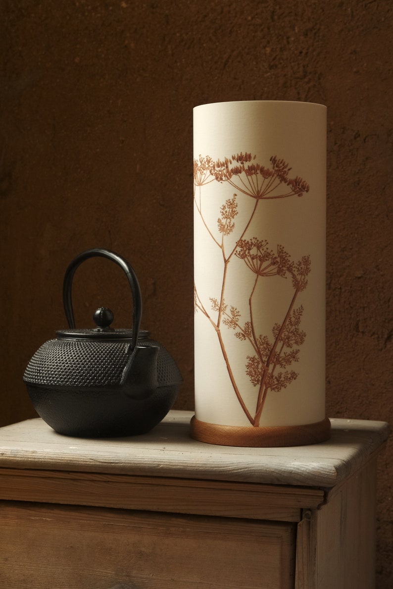 K&ouml;nnte beinhalten: Eine schwarze gusseiserne Teekanne mit Henkel steht auf einem Holztisch neben einer wei&szlig;en Lampe mit braunem Blumendesign.