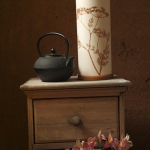 May include: A black cast iron teapot sits on a wooden table next to a white ceramic lamp with a dried flower design. A bouquet of pink flowers sits in front of the table.