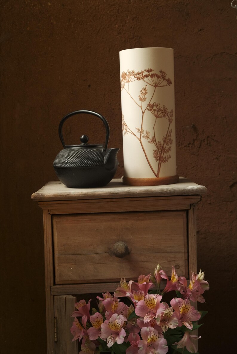 May include: A black cast iron teapot sits on a wooden table next to a white ceramic lamp with a dried flower design. A bouquet of pink flowers sits in front of the table.