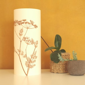 K&ouml;nnte beinhalten: Ein wei&szlig;er zylindrischer Lampenschirm mit braunem Blumendesign. Der Lampenschirm steht auf einer wei&szlig;en Oberfl&auml;che neben zwei Topfpflanzen. Eine Pflanze steht in einem geflochtenen Korb und die andere in einem grauen Topf.