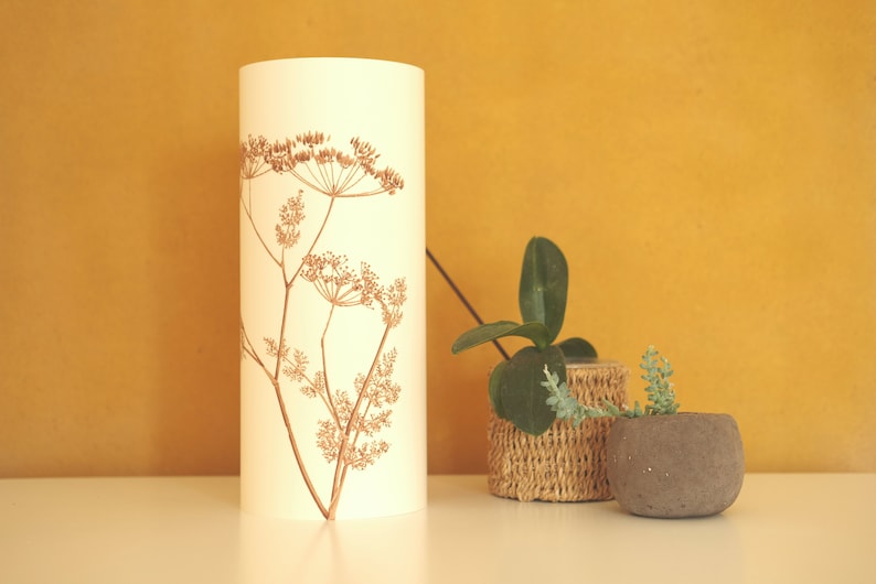 K&ouml;nnte beinhalten: Ein wei&szlig;er zylindrischer Lampenschirm mit braunem Blumendesign. Der Lampenschirm steht auf einer wei&szlig;en Oberfl&auml;che neben zwei Topfpflanzen. Eine Pflanze steht in einem geflochtenen Korb und die andere in einem grauen Topf.