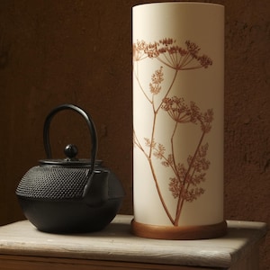 May include: A black cast iron teapot with a handle and spout sits on a wooden table next to a white cylindrical lamp with a brown floral design.