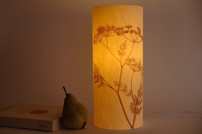 K&ouml;nnte beinhalten: Ein cremefarbener Lampenschirm mit einem gepressten Blumendesign einer zarten, braunen Wildblume. Der Lampenschirm ist beleuchtet und strahlt ein warmes Licht aus.