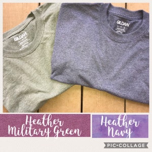 May include: Two Gildan brand t-shirts, one in heather military green and one in heather navy. The shirts are folded and laid out on a wooden surface.