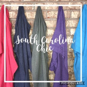 May include: Five different colored t-shirts hanging on a brick wall. The t-shirts are red, dark blue, gray, purple, and light blue. The text "South Carolina Chic" is written in white cursive letters across the front of the t-shirts.