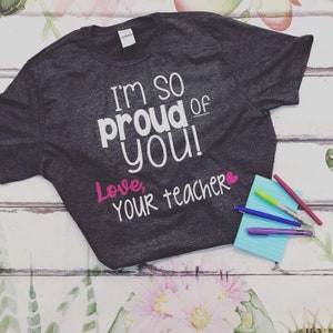 May include: A dark gray t-shirt with the text "I'm so proud of you! Love, your teacher" in white and pink lettering.