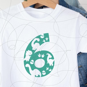 May include: White t-shirt featuring a teal number six design filled with dinosaur silhouettes, footprints, and eggs. The shirt is hanging on a wooden hanger against a denim jacket background. A fun and playful design for a child's birthday.