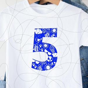Puede incluir: Camiseta blanca con un número 5 azul. El número está relleno con un patrón blanco de abejas, nubes, sol y otros diseños de temática veraniega.