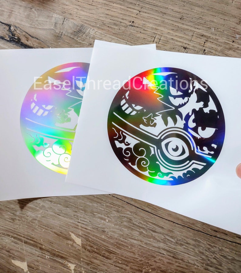 May include: Two iridescent vinyl stickers featuring a black and white design of a Pokeball with a Gengar silhouette inside. The stickers are on a white background.