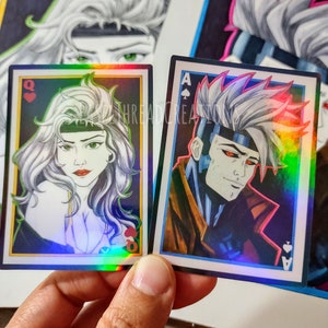 May include: Two holographic playing cards featuring illustrations of characters from the X-Men comics. The Queen of Hearts card features a portrait of Rogue, and the Ace of Spades card features a portrait of Gambit.