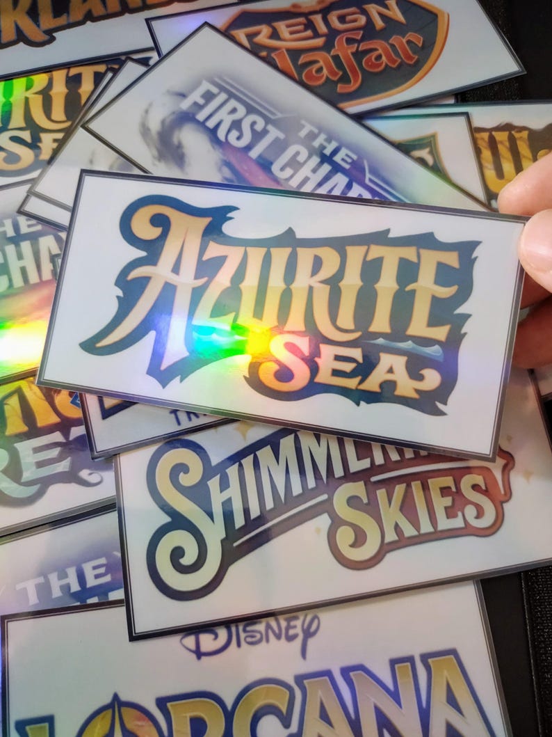May include: A stack of holographic stickers with various Disney movie titles, including "The First Chapter", "Reign of Jafar", "Azuritie Sea", "The Shimmering Skies", and "Disney Lorcana".