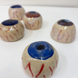 May include: A collection of ceramic eyeball sculptures. Each eyeball features a blue iris with a black pupil, set in a beige sclera with red vein detailing. The sculptures are designed to resemble realistic human eyes.