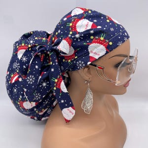 May include: A navy blue scrub cap with a festive Santa Claus and snowflake pattern. The cap is tied in a bow at the back and is worn with large, clear-lensed glasses with gold frames. The cap is a holiday-themed accessory.