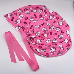 Könnte beinhalten: Rosa OP-Haube mit einem sich wiederholenden Muster aus Hello Kitty-Figuren, Schleifen und Äpfeln. Die Haube hat eine rosa Schleife zum Binden. Der Stoff ist mit dem Text "Hello Kitty" gemustert.