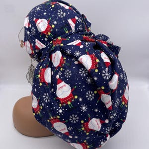 May include: A navy blue scrub cap featuring a repeating pattern of Santa Claus figures, snowflakes, and yellow dots. The Santa figures are dressed in red suits with white beards and hats. The cap is tied at the back.
