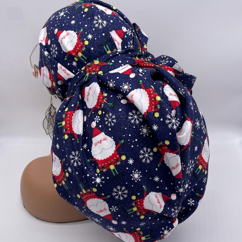May include: A navy blue scrub cap featuring a repeating pattern of Santa Claus figures, snowflakes, and yellow dots. The Santa figures are dressed in red suits with white beards and hats. The cap is tied at the back.