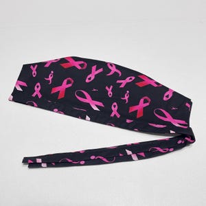 May include: A black surgical scrub cap featuring a pattern of pink ribbons. The ribbons are in various sizes and orientations, with some having a gradient effect. The cap has long ties for securing it.