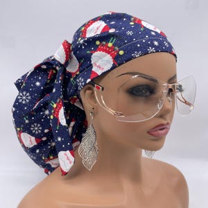 May include: A navy blue scrub cap with a Christmas Santa Claus print, tied in a bow. The cap is paired with gold-rimmed, clear-lens glasses and silver leaf earrings. The cap features a pattern of Santa Claus figures and snowflakes.